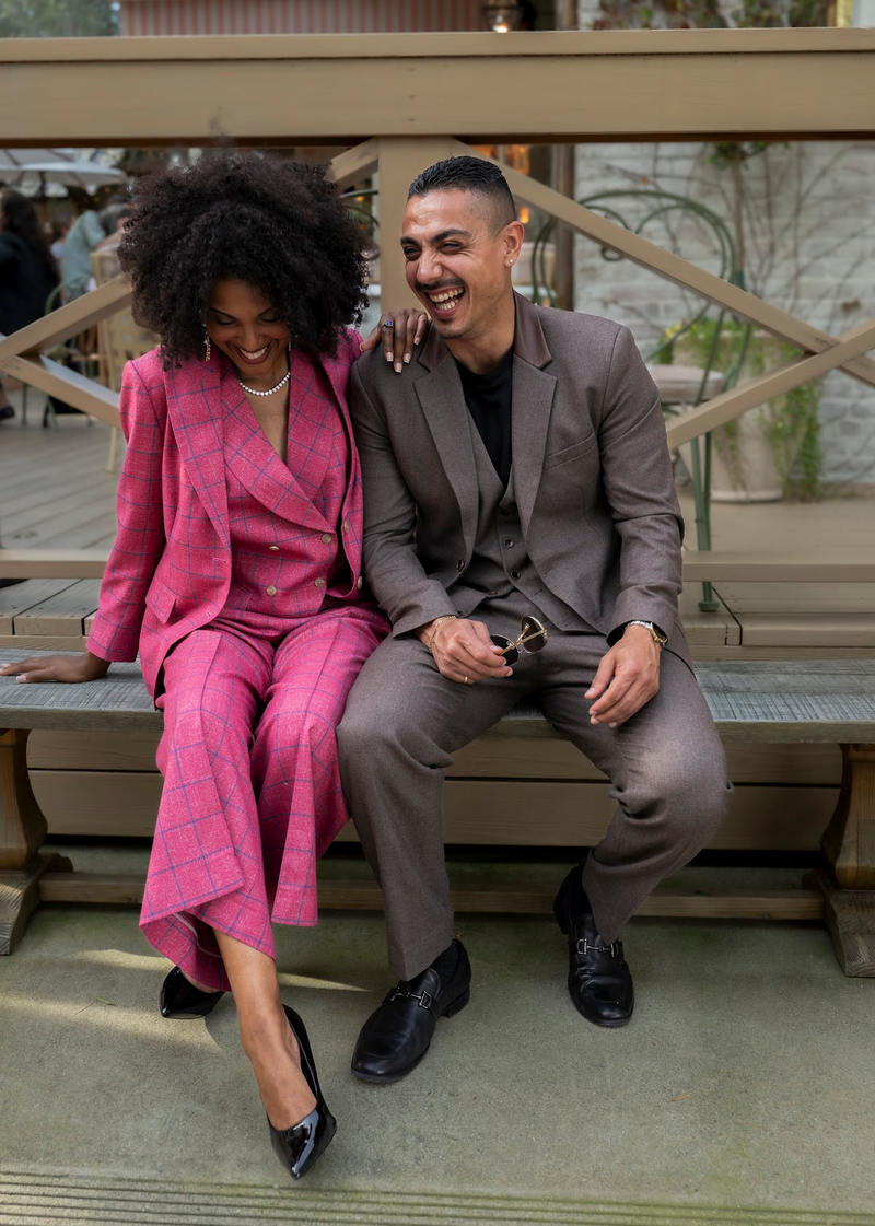 Coordinated bespoke suiting for power couples featuring a vibrant pink windowpane couture women's suit and a custom brown three-piece Seth Eden executive suit.