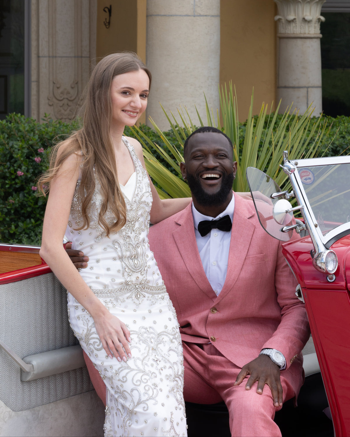 Coordinated luxury gala attire for power couples, featuring an intricately beaded white Nancy Vuu couture evening gown and a custom salmon pink Seth Eden tuxedo, posed by a classic red convertible at a private estate.