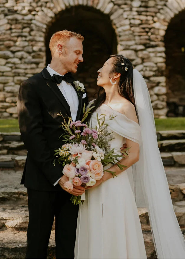 A smiling couple on their wedding day showcasing coordinated styling, featuring an elegant off-the-shoulder Nancy Vuu bespoke bridal gown with a long veil, and a classic black Seth Eden custom tuxedo.